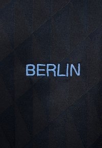 Μπλε κεντημένο κείμενο «BERLIN» σε μαύρο ύφασμα με διακριτικό γεωμετρικό μοτίβο τριγώνων στο φόντο.
