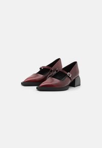 Burgundy leather shoes with a pointed toe, adjustable ankle strap, and a chunky black heel. Smooth texture and minimalistic design.