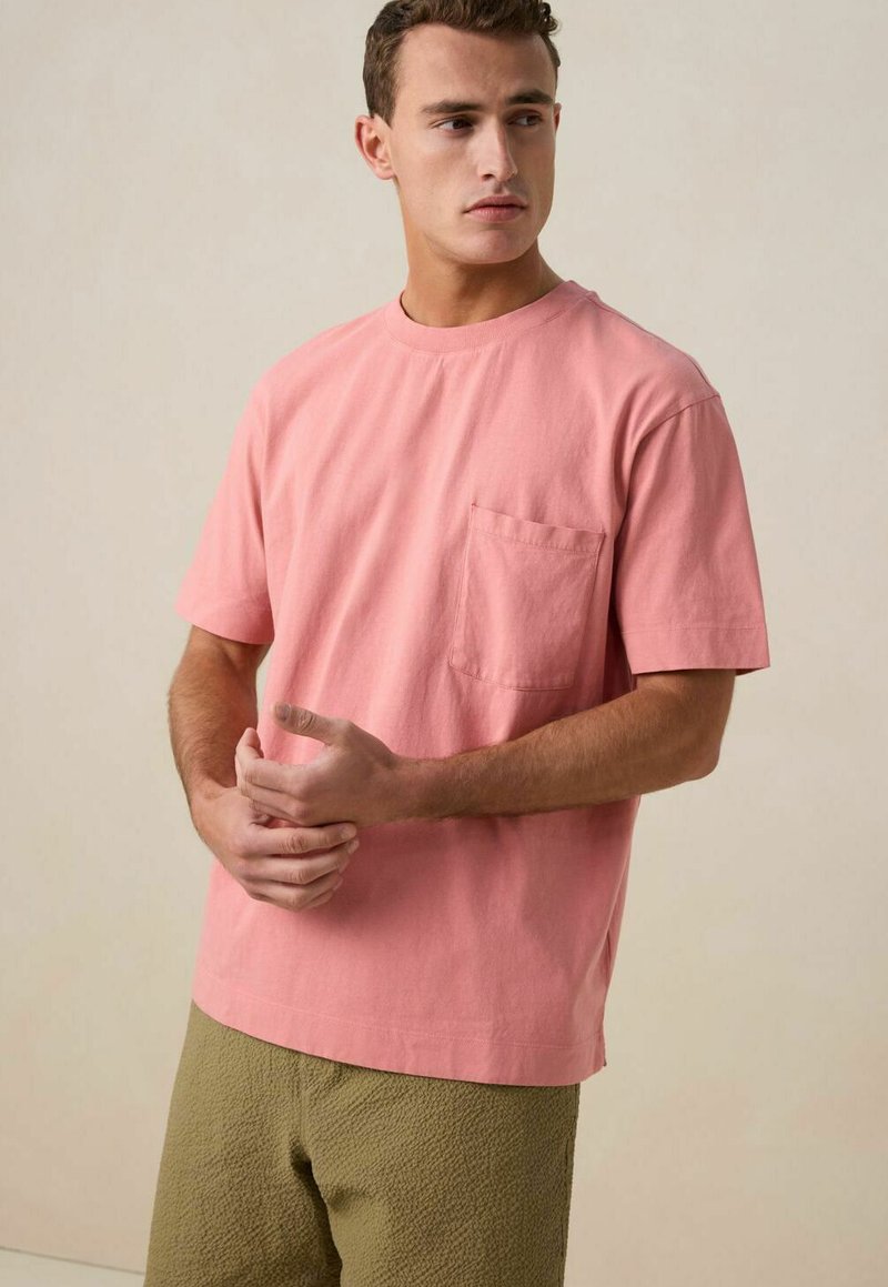 T-shirt en coton rose à manches courtes, col rond et poche poitrine. Porté avec un short vert texturé. Tissu lisse, coupe décontractée.