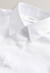 Biała koszula szkolna z kołnierzykiem, o gładkiej fakturze, z pięcioma guzikami i markową metką, ułożona w taki sposób, aby wyeksponować szczegóły kołnierzyka i guzików.