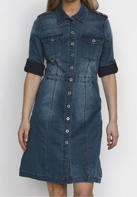 Denimmekko, jossa on kaulus, lyhyet käännetyt hihat, kaksi rinta-taskua, edessä nappikiinnitys ja istuva vyötärö, joka levenee halkiohameeseen.