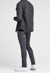 Mann in einer dunkelgrauen Jeansjacke, passenden engen Jeans und weißen Sneakers, der vor einem schlichten hellen Hintergrund steht und sich abkehrt.