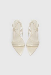 Sandales à talons hauts beige avec un bout pointu, présentant des lanières fines croisées et une finition translucide. Lanières réglables à la cheville incluses.