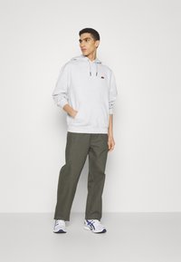 Sudadera gris claro con un bolsillo frontal, cordones negros y un pequeño logo. Combinada con pantalones verdes y zapatillas deportivas blancas.