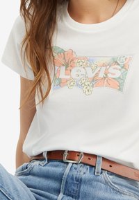 Camiseta blanca de algodón con un gráfico del logo floral de Levi's, combinada con unos vaqueros de denim claros y un cinturón de cuero marrón con hebilla de plata.