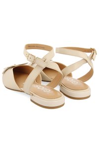 Sandalias de cuero beige con tiras cruzadas y hebilla en el tobillo, con suela plana y puntera redondeada pronunciada. Logo de la marca impreso en el interior.