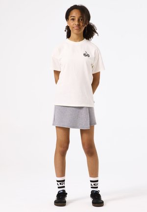 Weißes Baumwoll-T-Shirt mit schwarzem Kirschgrafik, kombiniert mit einem gefalteten grauen Rock, schwarzen Sneakers und gemusterten schwarz-weißen Socken.