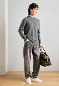 Pulover gri din lână, blugi gri estompați, adidași albi și o geantă verde măslin cu accente maro. Modelul stă pe un podea din lemn, în fața unui perete minimalist.