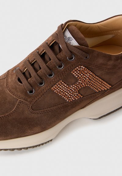 Sneaker in suede marrone con lacci marroni, suola beige, colletto imbottito e un logo decorativo arancione di strass a forma di "H" sul lato.