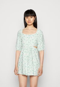 Vestido floral curto em verde claro e branco, apresentando mangas bufantes, decote quadrado, detalhes recortados e uma cintura franzida.