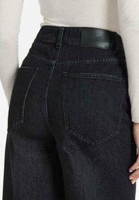Donkerdenim high-waisted jeans hebben een gebogen achterzakontwerp, een zwart leren label op de tailleband en een gestructureerde stofafwerking.