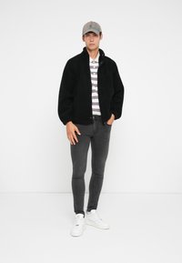 Svart fleecejakke, hvit og stripete polo, grå slim-fit jeans og hvite joggesko. Modellen har på seg en lys grå caps. Enkel bakgrunn.