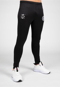 Black athletic pants with a slim fit, made of stretchy material. Features white logo graphics and a waistband with elastic.