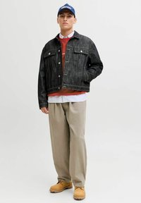 Jeansowa kurtka w ciemnym odcieniu z przednimi kieszeniami, nałożona na czerwoną sweter, białą koszulę z kołnierzykiem i beżowe spodnie. Ciemnobrązowe buty dopełniają całość.