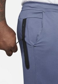 Blauwe sportbroek voor heren met een zijzak met ritssluiting en zwarte hardware, gemaakt van textuurstof. Close-up van de zak en de tailleband.