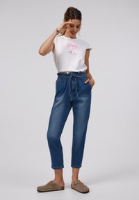 Witte t-shirt met een roze satijnen strikdetail, gecombineerd met hoog-gepositioneerde blauwe denim broeken met een elastische tailleband en casual instappers.