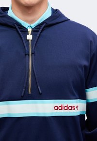 Άνδρας που φορά μπλε μπουφάν με κουκούλα με ανοιχτό μπλε και λευκή ρίγα στο στήθος και το μανίκι, κόκκινο λογότυπο της Adidas στη ρίγα του στήθους.