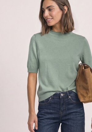 Femme portant un pull à manches courtes vert clair et un jean bleu foncé, tenant un sac cabas en daim marron, regardant sur le côté et souriant.