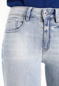 Svijetloplavi traperice ravnog kroja, s visokim strukom, prednjim džepovima i suptilnim izbljeđivanjem za efekt iznošenosti. Srebrni metalni detalji.