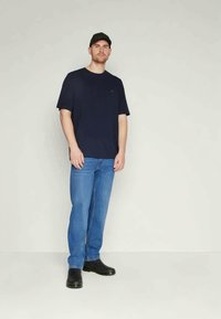 Tricou din bumbac bleu marin cu mâneci scurte, asortat cu blugi din denim bleu deschis. Modelul poartă pantofi negri și o șapcă neagră.