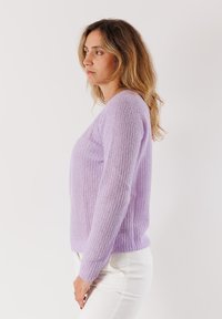 Donna con capelli castani chiari e mossi, che indossa un maglione di lana lilla e pantaloni bianchi, in piedi di profilo su uno sfondo bianco semplice.