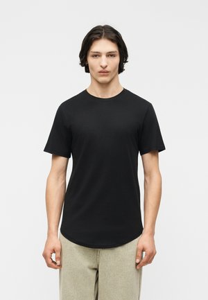 Giovane uomo con capelli castano scuro di media lunghezza, indossa una maglietta nera a maniche corte e pantaloni beige chiaro, in piedi contro uno sfondo bianco.