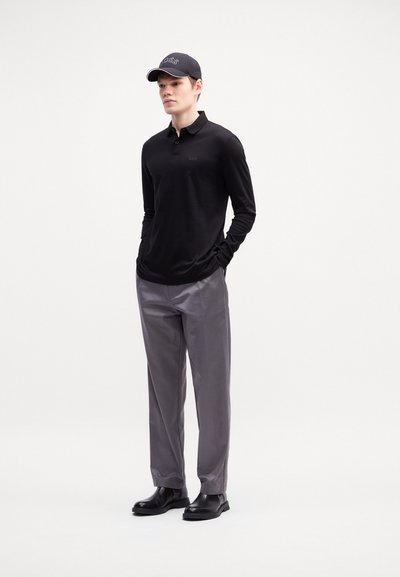 Polo noir à manches longues avec un logo discret sur la poitrine, associé à un pantalon gris et des chaussures en cuir noires, complété par une casquette noire.