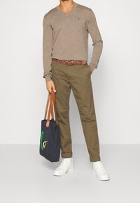 Světle hnědý svetr s výstřihem do V, olivově zelené chinos kalhoty, hnědý tkaný opasek, bílé tenisky a námořnicky modrá taška přes rameno s multicolor grafikou a hnědými popruhy.