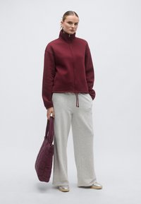 Vīnsarkans džemperis ar rāvējslēdzēju, pelēkas plata auguma sporta bikses un vīnsarkana krokaina tote soma. Metāliskas sudraba krāsas apavi ar teksturētu, ikdienišķu izskatu.