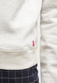 Svetle šedá mikina s žebrovaným lemem a manžetami, s červenou štítkem Levi's na straně. Měkká textura s volným střihem.