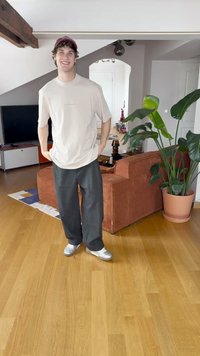 T-shirt bege claro, calças pretas largas e sapatilhas prateadas. Ambiente interior com um sofá de veludo cotelê castanho e uma grande planta verde.