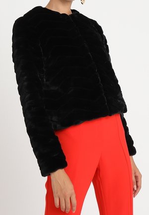 Woman wearing black textured faux fur jacket and bright red pants, standing against a plain white background.