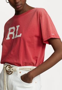 Röd bomullst-shirt med vit "RL" logotyp, korta ärmar och ribbad rundad halsringning. Matchas med vita denim och ett knutet repbälte.