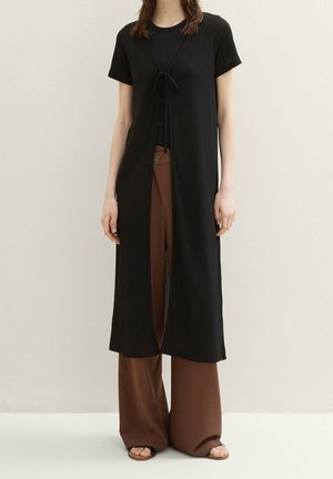 Kobieta w czarnej, krótkorękawowej, długiej kamizelce zawiązanej na klatce piersiowej, czarnym topie, brązowych szerokich spodniach i sandałach na odkryte palce, stojąca.