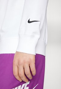 Mano descansando a un lado, vistiendo un sudadera blanca de Nike con un logo negro en la manga y pantalones cortos morados de Nike con un logo blanco.