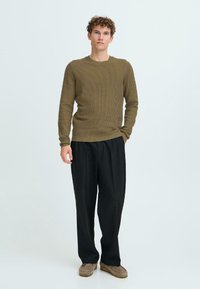 Pulover ribbed de culoare verde măsliniu, cu guler rotund, purtat cu pantaloni negri largi și pantofi de culoare maro deschis, pe un fundal neutru.