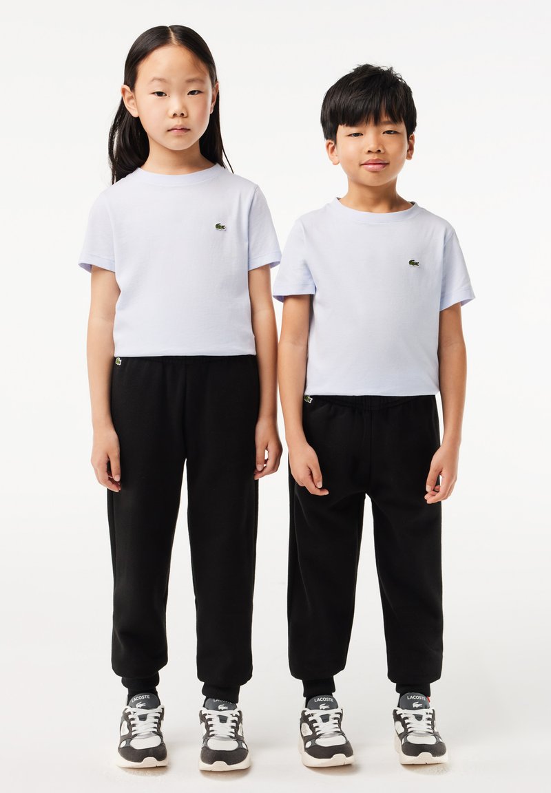 Helllila Kurzarm-T-Shirts mit grünem Logo, kombiniert mit schwarzen schmal geschnittenen Jogginghosen und grauen Sneakers. Einfaches und lässiges Design.