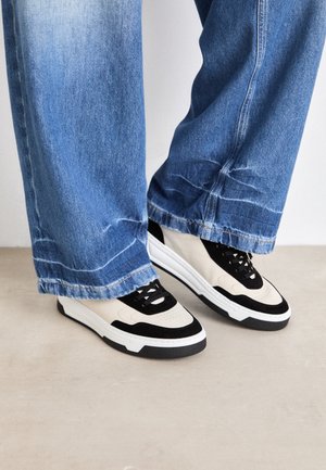 Osoba ubrana w szerokie niebieskie dżinsy i czarno-białe tenisówki stojąca na jasnej podłodze na tle gładkiej ściany.