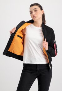 Zwarte bomberjack met gestructureerde buitenkant, oranje voering en een ritszak op de mouw. Gekozen boven een wit shirt en zwarte broek.
