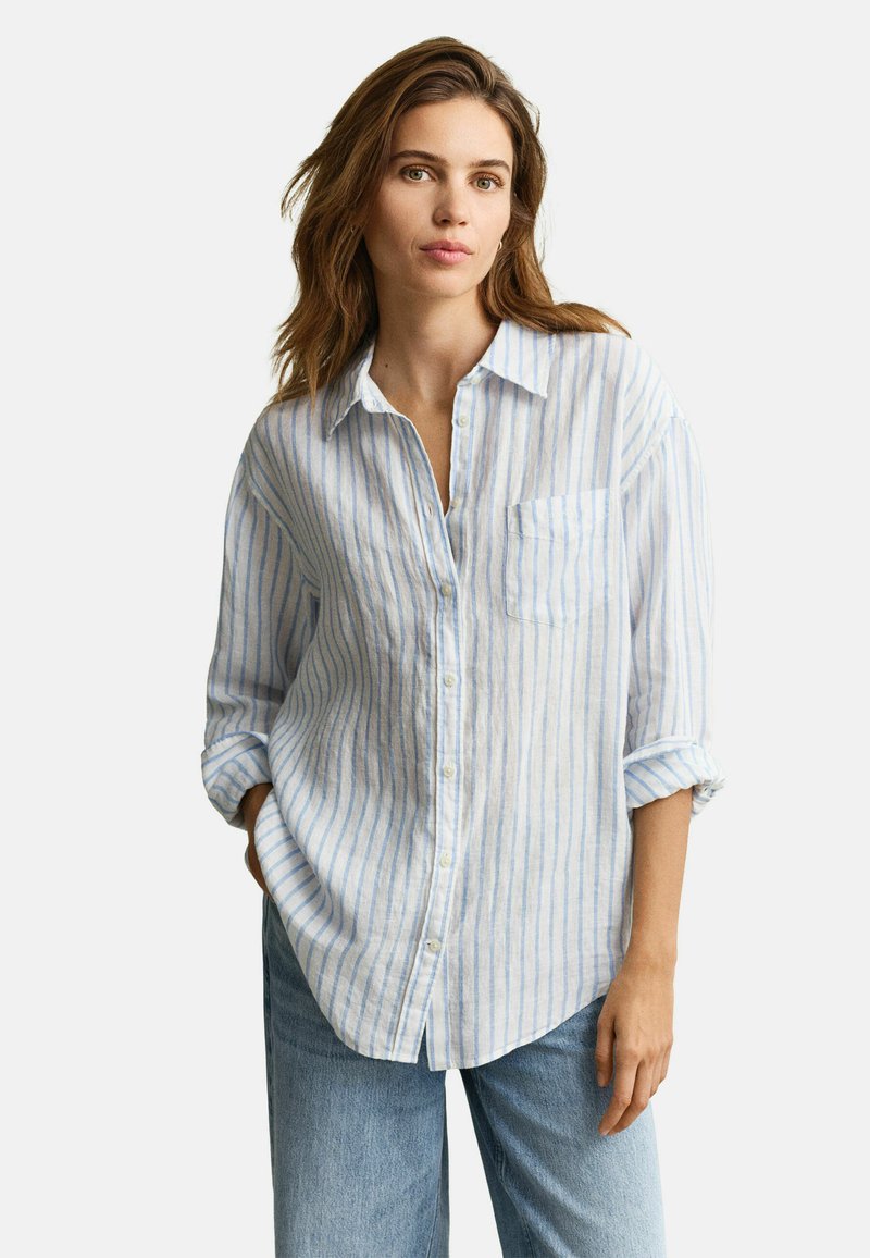 Mujer con cabello castaño claro que lleva una camisa blanca de botones con rayas verticales azules y jeans azul claros, de pie con una mano en el bolsillo.