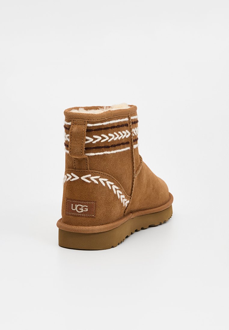 Stivale alla caviglia in suede marrone con cuciture decorative bianche, fodera in shearling, suola piatta in gomma e etichetta con logo UGG sul retro.