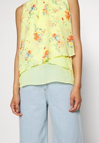 Blusa sin mangas floral en amarillo brillante, con diseño en capas, tela ligera y varios colores que incluyen flores en naranja y azul.
