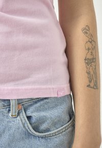 Enganliegendes pinkes Tanktop aus weichem Material, am Taillenbund gesäumt, mit einem kleinen Logo-Patch, kombiniert mit hellblauen Jeans.