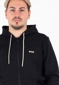 Schwarze Zip-Hoodie aus Baumwollmischung, mit einer Kapuze mit Kordelzug und einem kleinen weißen "BOSS"-Logo auf der Brust, mit beigen Kordelzügen.