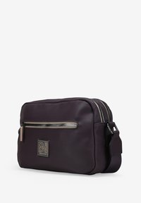 Bolso de bandolera de nylon morado con un bolsillo frontal con cremallera, herrajes en plata, parche con logo y una correa ajustable. Diseño compacto y rectangular.