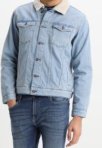 Vaaleansininen farkkutakki, jossa on sherpa-kaulus, napitus, kaksi rintataskua ja sopiva malli. Päällä tummansinisiä farkkuja.