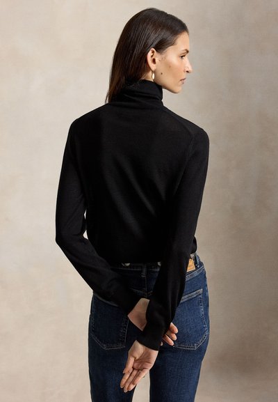Femme aux cheveux foncés porte un pull à col roulé noir et un jean bleu, debout de profil, les mains jointes dans le dos.