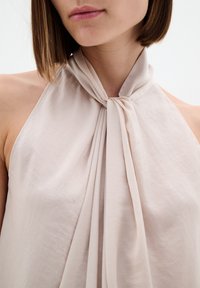 Světlobéžový top na zavazování za krkem s volným střihem, vyrobený z jemného texturovaného materiálu a s něžným splýváním kolem krku.