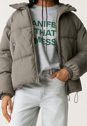 Doudoune grise avec capuche, texture matelassée, fermetures éclair, associée à un t-shirt graphique gris clair et un jean bleu clair.