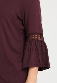 Burgundianpunainen paita, jossa on kolmivartiset kellohihat. Hihansuun reuna on pitsimäisellä leikkauskuviolla, valmistettu pehmeästä, kevyestä kankaasta.